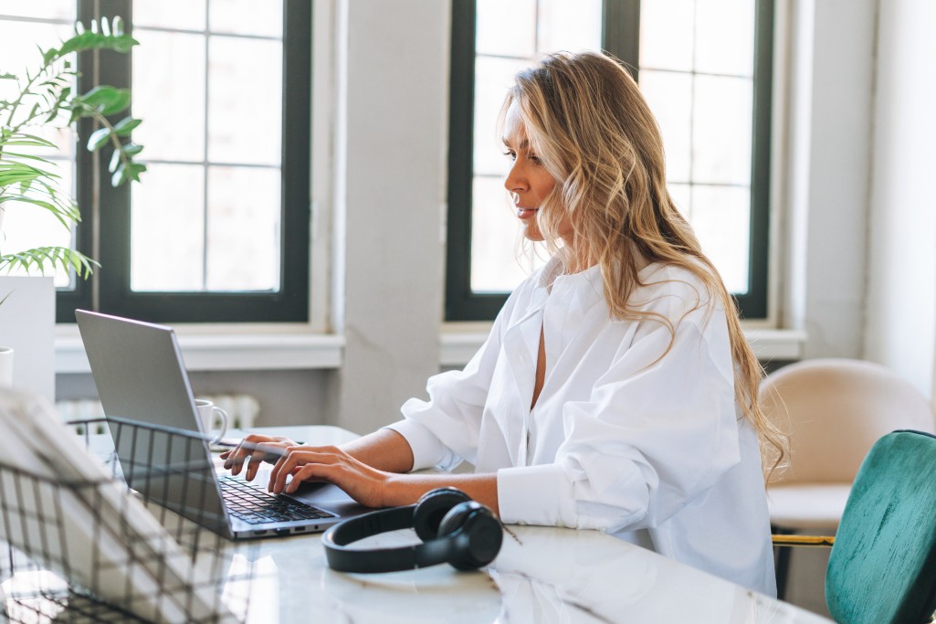 young-smiling-blonde-woman-with-long-hair-in-stylish-wite-shirt-working-at-laptop-in-the-bright